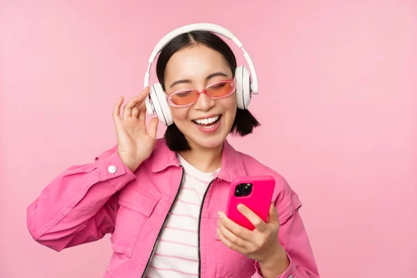 Elegante chica asiática bailando con smartphone, escuchando música en auriculares en la aplicación del teléfono móvil, sonriendo y riendo, posando sobre fondo rosa — Foto de Stock