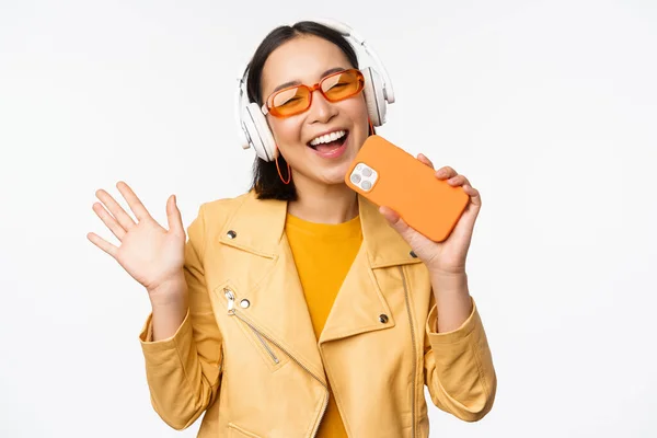 Szczęśliwa stylowa Azjatka w okularach przeciwsłonecznych, słuchająca muzyki w słuchawkach, trzymająca smartfona, śpiewająca i tańcząca, stojąca na białym tle — Zdjęcie stockowe