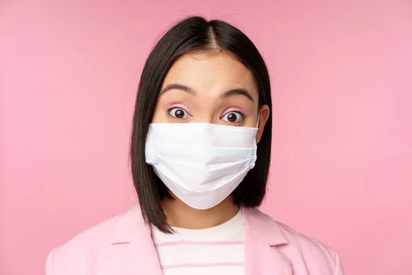 Close-up retrato de asiático empresária em médico máscara facial, olhando surpreendido, de pé em terno sobre rosa fundo — Fotografia de Stock