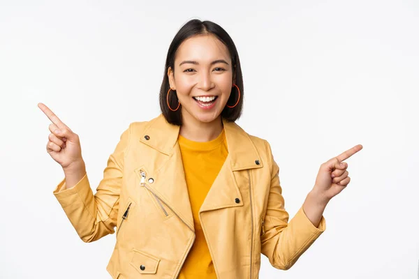 Glad vacker asiatisk flicka pekar finger vänster och höger, visar banner logotyp, visar försäljning, står i gul jacka över vit bakgrund — Stockfoto