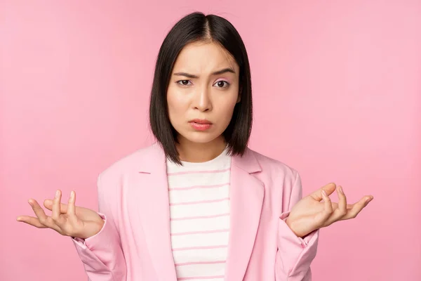 Retrato de joven mujer de negocios asiática, vendedora encogiéndose de hombros y buscando confundido, despistado de smth, de pie sobre fondo rosa —  Fotos de Stock