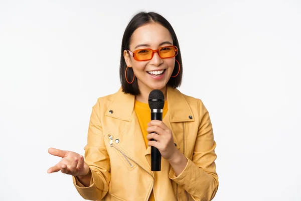 Retrato de hermosa mujer asiática en gafas de sol, chica elegante cantando, dando discurso con micrófono, sosteniendo micrófono y sonriendo, de pie sobre fondo blanco — Foto de Stock