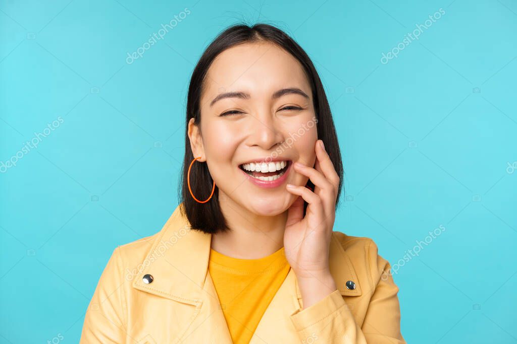 Close up portrait of natural asian girl laughing, smiling and looking happy, standing over blue background. Copy space