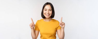 Güzel Japon kızın portresi gülümsüyor, parmaklarını havaya kaldırıyor, reklamını yapıyor, sarı tişörtle beyaz arka planda dikiliyor.