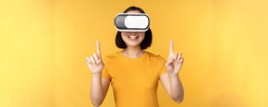 VR 'da oynayan Asyalı genç bir kadın, sanal gerçeklik gözlüğü takıyor, stüdyo arka planında sarı tişört giyiyor.