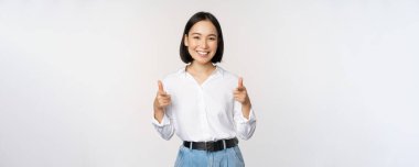 Tebrik ederim. Gülümseyen çekici Asyalı kadın, iş kadını mutlu bir yüzle kamerayı işaret ediyor, iltifat ediyor, davet ediyor, beyaz arka planda duruyor.