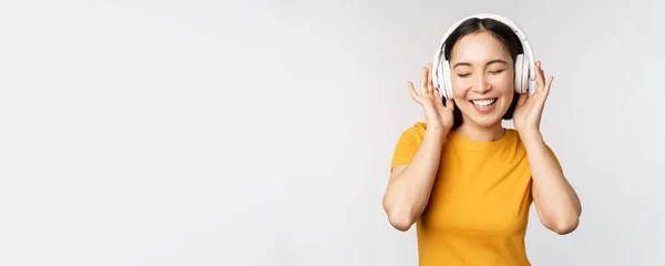 Feliz chica asiática bailando, escuchando música en los auriculares y sonriendo, de pie en camiseta amarilla sobre fondo blanco — Foto de Stock