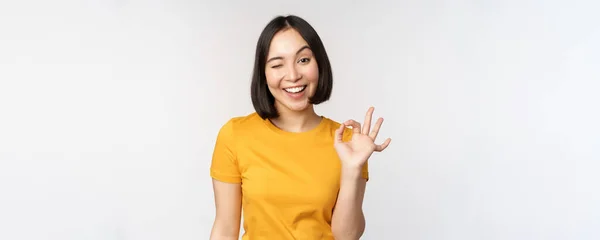 Hermosa mujer joven mostrando signo bien, sonriendo complacido, recomendando smth, aprobar, como producto, de pie en camiseta amarilla sobre fondo blanco —  Fotos de Stock