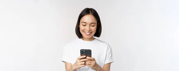 Portrait of smiling asian woman using mobile phone, chatting, texting message, standing in tshirt over white background — Stock Photo, Image