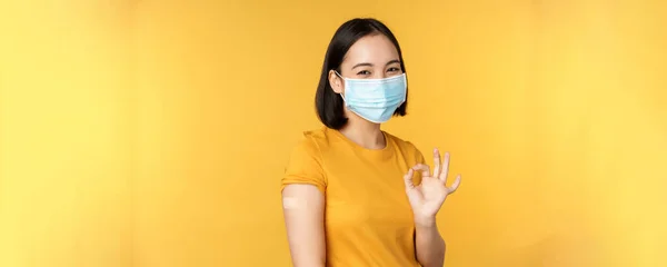 Εμβολιασμός από την έννοια covid και την υγεία. Χαρούμενη Ασιάτισσα που δείχνει καλά, φοράει ιατρική μάσκα, επίδεσμο στον ώμο, έχει εμβόλιο για τον ιό της στέψης, κίτρινο φόντο. — Φωτογραφία Αρχείου
