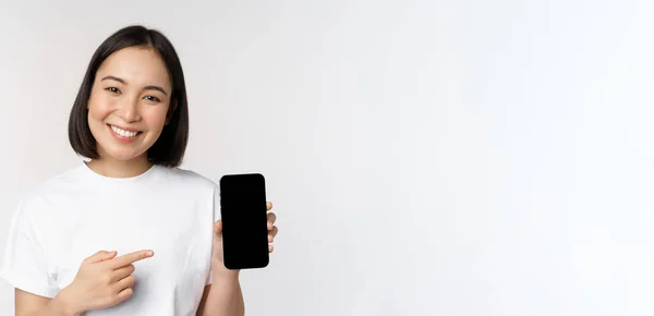 Leende asiatisk kvinna pekar finger på smartphone skärm, visar applikationsgränssnitt, mobiltelefon webbplats, stående över vit bakgrund — Stockfoto