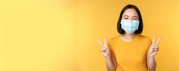 Positiv asiatisk kvinna ler, bär medicinsk ansiktsmask från covid-19 under pandemi, visar fred v-tecken gest, gul bakgrund — Stockfoto