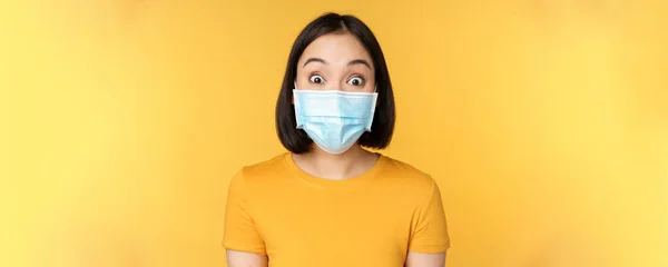 Covid-19 and medical concept. Close up portrait of asian woman in face mask, looking surprised and amazed at news, standing over yellow background — Stock Photo, Image