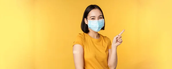 Vaccinatie van covid en gezondheid concept. Afbeelding van glimlachend Koreaans meisje in medisch gezichtsmasker, pleister op de schouder, wijzende vinger naar banner reclame, gele achtergrond — Stockfoto