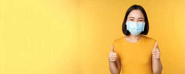 Sorrindo mulher asiática em máscara facial médica, mostrando polegares para cima aprovação, como e recomendo smth, de pé sobre fundo amarelo — Fotografia de Stock