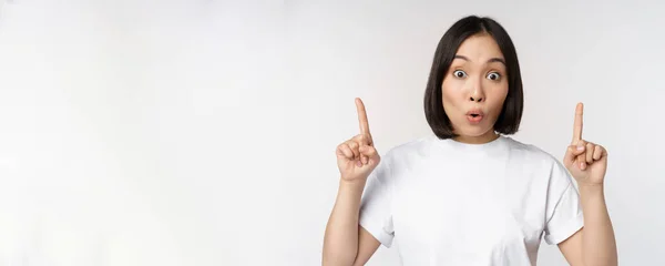 Портрет захопленої молодої жінки, азіатська дівчина посміхається вказуючи пальці вгору, показуючи рекламу вгору, стоячи на білому тлі — стокове фото