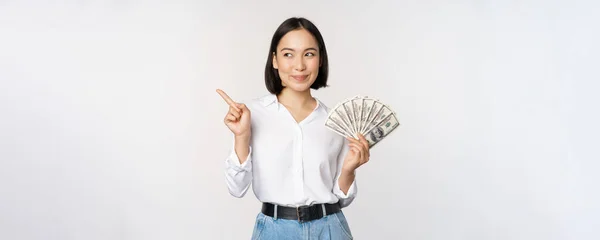 Tersenyum wanita muda Asia modern, menunjuk pada iklan banner, memegang uang tunai dolar, berdiri di atas latar belakang putih — Stok Foto