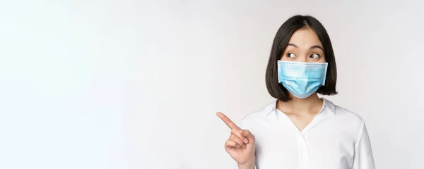 Portret van een leuke Aziatische vrouw met medisch gezichtsmasker, bescherming tegen het coronavirus, wijsvinger links en geïntrigeerd kijkend naar lege kopieerruimte, witte achtergrond — Stockfoto