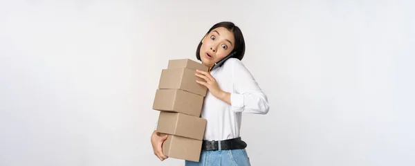 Junge asiatische Geschäftsfrau nimmt Anruf entgegen, telefoniert mit dem Handy, während sie vor weißem Hintergrund stapelweise Kartons mit Bestellungen trägt — Stockfoto