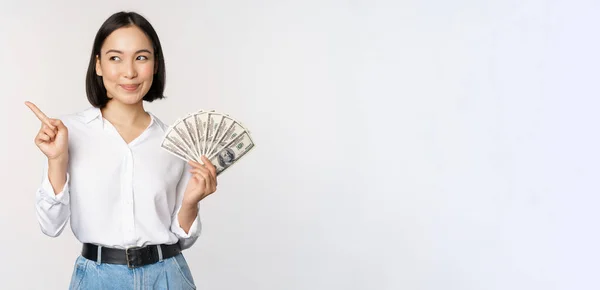 笑顔の若い現代アジアの女性は、バナー広告を指して、現金のお金のドルを保持し、白い背景の上に立って — ストック写真