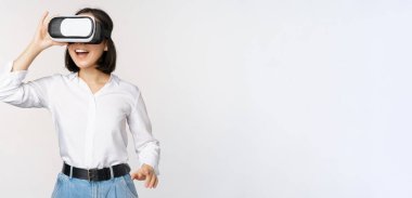 Heyecanlı genç kadın gözlükleriyle sanal gerçekliğe giriyor. Beyaz arka planda duran, video kulaklık kullanan Asyalı bir kız.