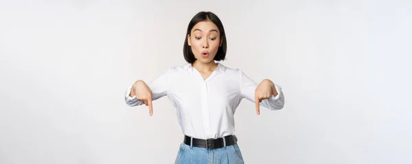 Překvapený mladý asijský studentka, ukazuje prsty dolů a dívá se s ohromeným, ohromený výraz obličeje, stojící nad bílým pozadím — Stock fotografie