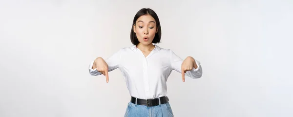 驚くべき若いアジアの女性学生,指を下に指して、驚きを見て,印象的な顔の表情,白い背景の上に立って — ストック写真