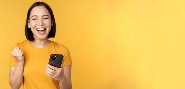 Glädjande asiatisk kvinna firar, håller mobiltelefon, vinna, uppnå mål på smartphone, står över gul bakgrund — Stockfoto