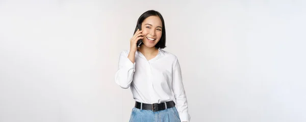 Ystävällinen hymyilevä aasialainen nainen puhuu puhelimessa, tyttö päivystää, pitää älypuhelinta ja nauraa, puhuu, seisoo valkoisen taustan yläpuolella — kuvapankkivalokuva
