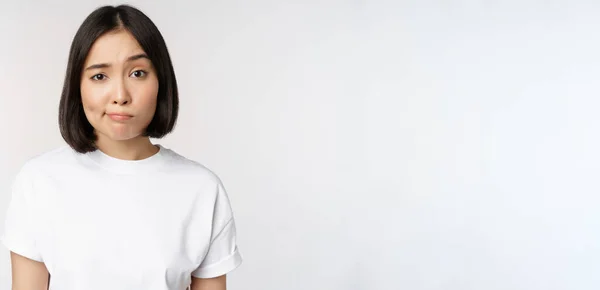 Image d'une jeune femme brune asiatique en t-shirt blanc, souriant et regardant avec doute, hésitant, debout sur fond blanc — Photo