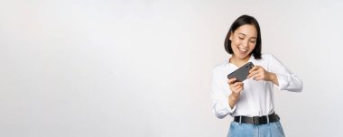 Genç Koreli kadın, akıllı telefondan mobil video oyunu oynayan Asyalı kız, yatay telefon ekranına bakıyor, beyaz arka planda duruyor.