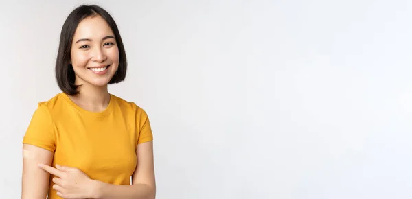 Vaccinationskampanj mot Covid 19. Leende asiatisk flicka pekar på hennes band stöd på axeln, rekommendera att få vaccin från omicron coronavirus, vit bakgrund — Stockfoto