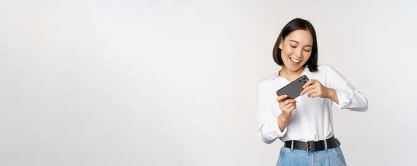 Mujer coreana joven, chica asiática jugando videojuego móvil en el teléfono inteligente, mirando la pantalla del teléfono horizontal, de pie sobre fondo blanco — Foto de Stock