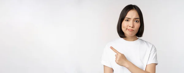 Escéptica chica asiática en camiseta blanca, apuntando al producto o logotipo con mueca decepcionada, disgusto y quejarse en smth, de pie sobre fondo blanco — Foto de Stock