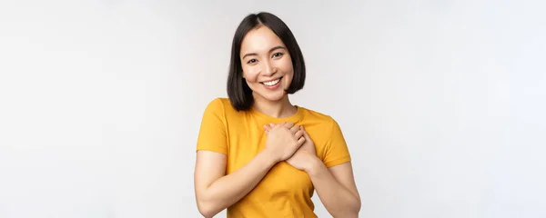 낭만적 인 동양 여자 친구, 가슴을 잡고, 주의와 부드러움을 가지고 미소를 짓고, 하얀 배경 위에 노란 티셔츠를 입고 서 있는 모습 — 스톡 사진