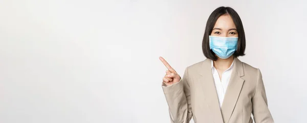 Коронавирус и концепция работы. Портрет женщины в маске медицинского лица, указывая пальцем влево, показывая логотип или баннер, реклама, белый фон — стоковое фото