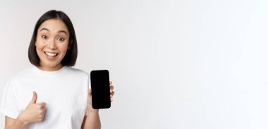 Hevesli genç kadın başparmağını kaldırıyor ve cep telefonu ekranında beyaz arka planda tişörtle dikiliyor.