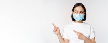 Tıbbi maske takan genç Koreli kadın solu işaret ediyor ve logoya bakıyor, reklam ya da afiş gösteriyor, beyaz arka planda duruyor.