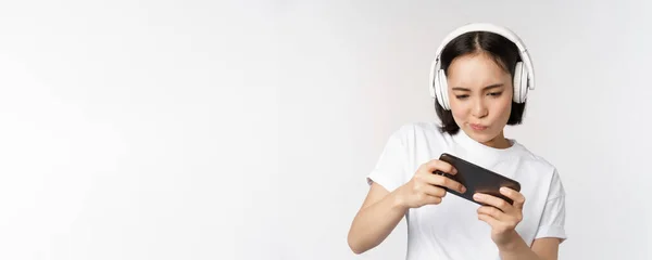 Mujer asiática feliz en los auriculares, mirando el teléfono inteligente, viendo vídeo en el teléfono móvil y sonriendo, de pie sobre fondo blanco — Foto de Stock