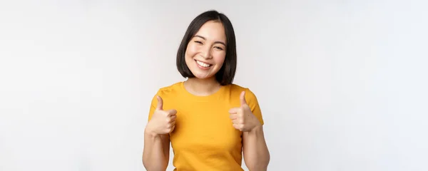 Bonito sorrindo asiático modelo feminino, mostrando polegares para cima e olhando satisfeito, recomendando, expressar feedback positivo, de pé sobre fundo branco — Fotografia de Stock