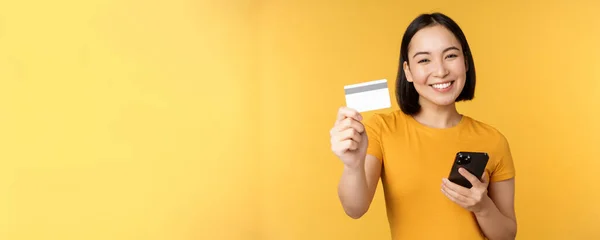 Joyful menina asiática sorrindo, mostrando cartão de crédito e smartphone, recomendando telefone celular bancário, de pé contra fundo amarelo — Fotografia de Stock