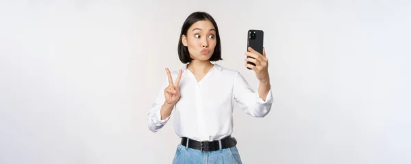 Divertida chica asiática mostrando paz, v-signo en la cámara del teléfono inteligente, posando para la foto, tomando fotos con aplicación móvil, de pie sobre fondo blanco — Foto de Stock