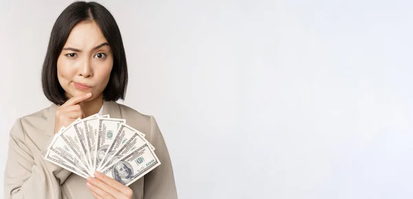 Образ азиатской корпоративной женщины, счастливой деловой женщины, показывающей деньги, наличные доллары и думающей, стоящей в костюме на белом фоне — стоковое фото