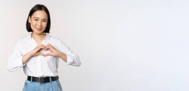 Aşk jesti. Güzel Asyalı genç kadın, kalp hareketi yapıyor ve gülümsüyor, özen ve şefkat gösteriyor, beyaz arka planda duruyor.