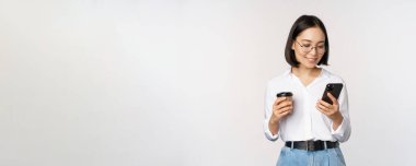Modern Asyalı kadının cep telefonuna baktığı, paket kahve içtiği, gözlük taktığı, akıllı telefon uygulaması kullandığı, beyaz arka planda durduğu bir resim.