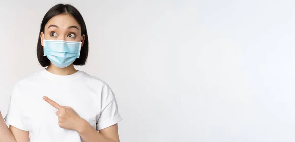 Молодая корейская женщина в медицинской маске для лица указывает пальцами влево и смотрит на логотип, показывая рекламу или баннер, стоя на белом фоне — стоковое фото