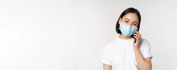 Gesundheit und Covid-19-Konzept. Lächelndes asiatisches Mädchen mit medizinischer Gesichtsmaske, telefonierend, Handy-Anruf entgegennehmen, vor weißem Hintergrund stehen — Stockfoto