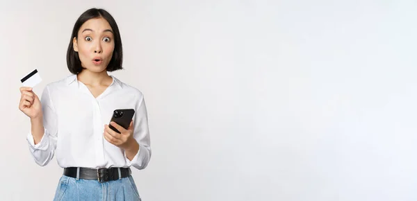Concepto de compras online. Imagen de chica asiática sorprendida, sosteniendo la tarjeta de crédito y el teléfono inteligente, mirando sorprendido en la incredulidad de la cámara, fondo blanco —  Fotos de Stock