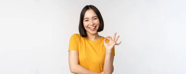 Schöne junge Frau zeigt Okay Zeichen, lächelt zufrieden, empfiehlt etw., genehmigen, wie Produkt, steht in gelbem T-Shirt über weißem Hintergrund — Stockfoto