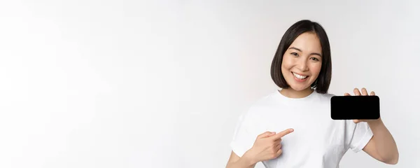 Retrato de mujer coreana sonriente apuntando con el dedo a la pantalla del teléfono móvil, mostrando la pantalla horizontal del teléfono inteligente, recomendando sitio web o tienda en línea, fondo blanco — Foto de Stock